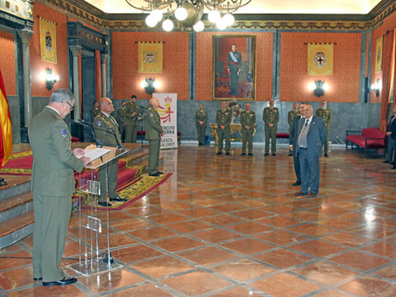 Personalidades militares en una ceremonia formal en una sala con mosaicos de azulejos rojos y amarillos, con banderas y retratos en el fondo.
