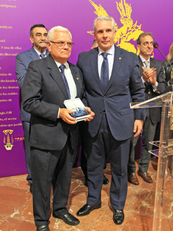 Dos hombres en trajes de negocios sostienen un libro mientras otros aplauden en el fondo. El fondo tiene un logotipo dorado y una pared de color púrpura.