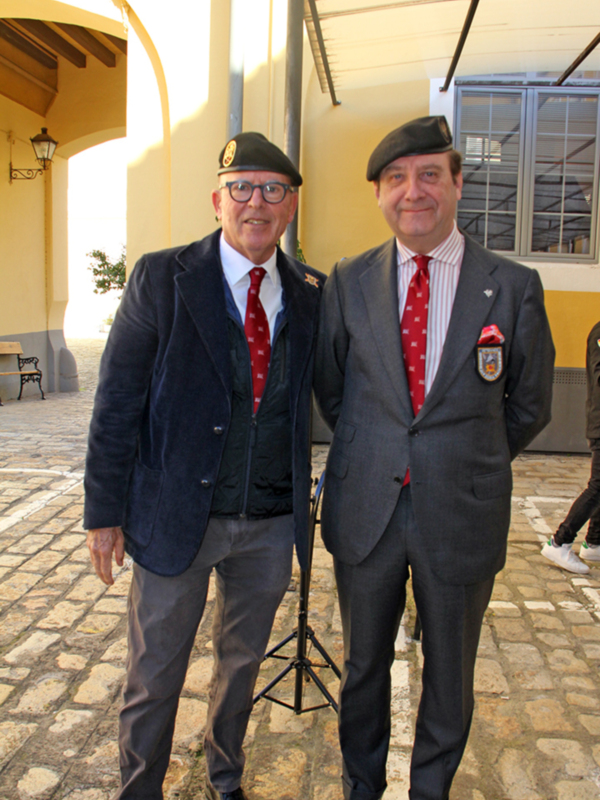 Dos hombres en trajes formales posan para una foto en un entorno urbano con una estructura de techo y una ventana en el fondo.