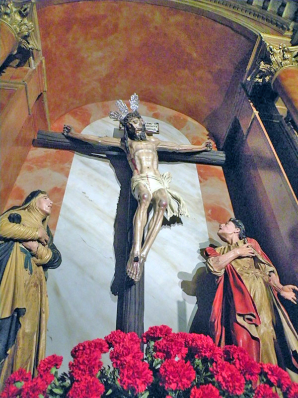 Representación escultórica del Cristo crucificado con dos figuras de fieles en adoración, rodeado de flores rojas.
