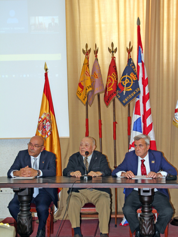 Tres hombres sentados en un podio con banderas y proyección de fondo.