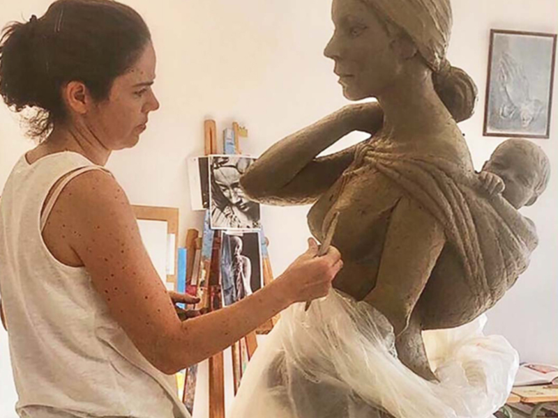 Artista escultora trabajando en una figura de mujer con bebé en brazos, en un taller de arte.