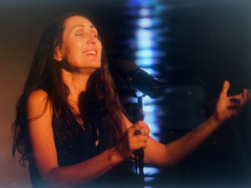 Una mujer con cabello largo y oscuro está cantando con emoción, sosteniendo un micrófono en una escena de concierto.