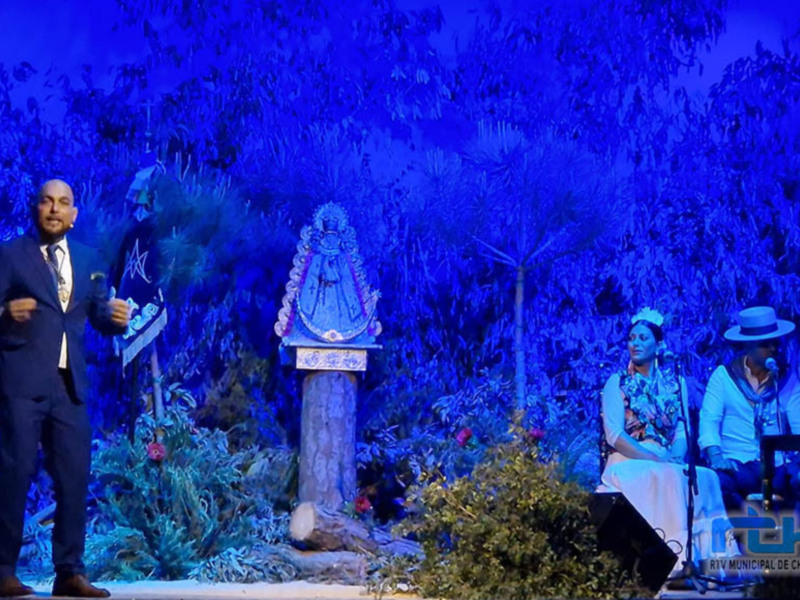 Un hombre en traje negro se dirige a una audiencia en un escenario con una estatua de la Virgen María en el centro, rodeada de vegetación y luces azules.