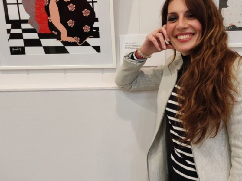 Una mujer sonríe en una galería de arte, detrás de ella se ve una obra abstracta con patrones geométricos y rosas rojas.