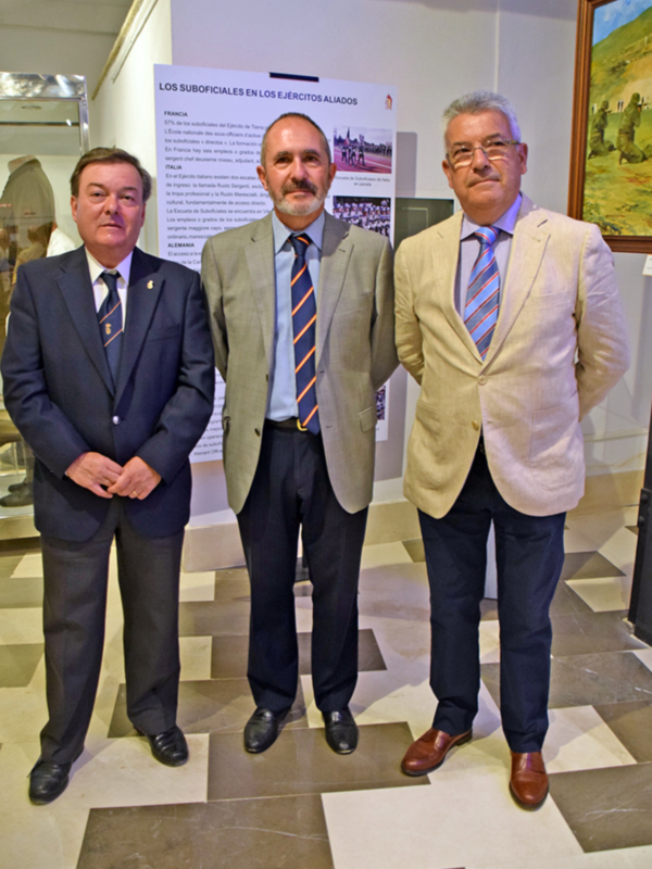 Dos hombres en trajes posan frente a un póster informativo sobre los suboficiales en los Ejércitos Aliados, con una imagen de un paisaje en el fondo.