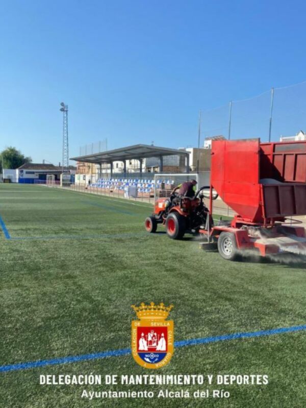 Tractor trabajando en el mantenimiento de un campo de fútbol artificial en Alcalá del Río, Sevilla. El tractor rojo está aplicando material en el césped para mejorar su calidad y durabilidad.