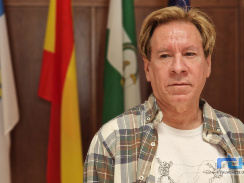 Un hombre con una camisa a cuadros y una camiseta con un dibujo en el pecho, frente a banderas.