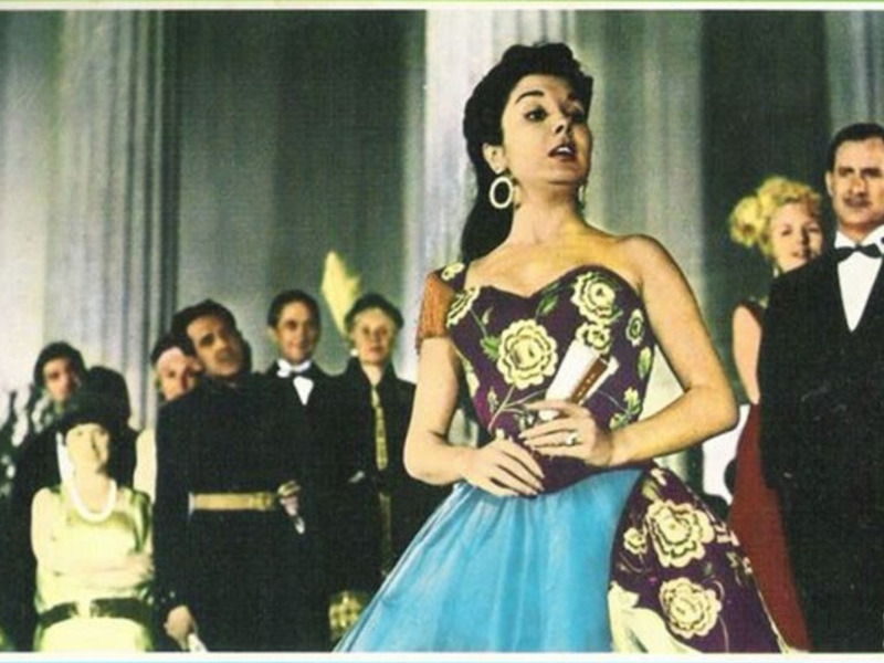 Escena de una película clásica con una mujer en un vestido floral y un grupo de personas en trajes de época en el fondo.