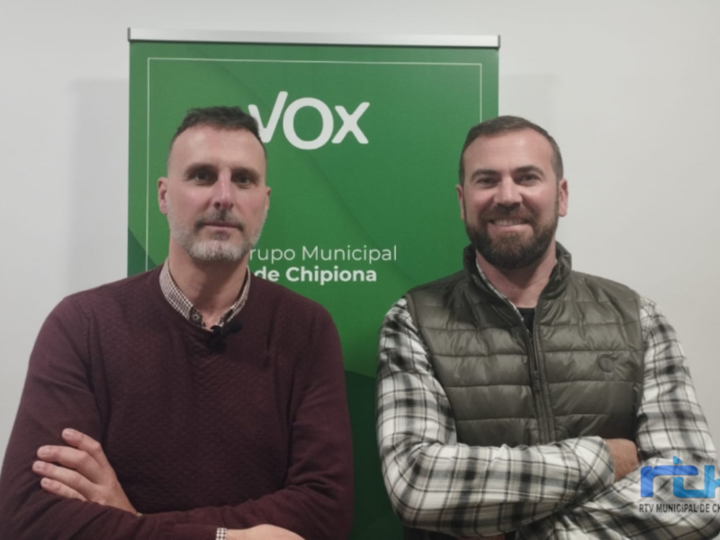Dos personas posando frente a un banner verde con el texto 'Grupo Municipal de Chipiona' y 'RTV Municipal de Chipiona'. Ambas tienen los brazos cruzados y sonríen.