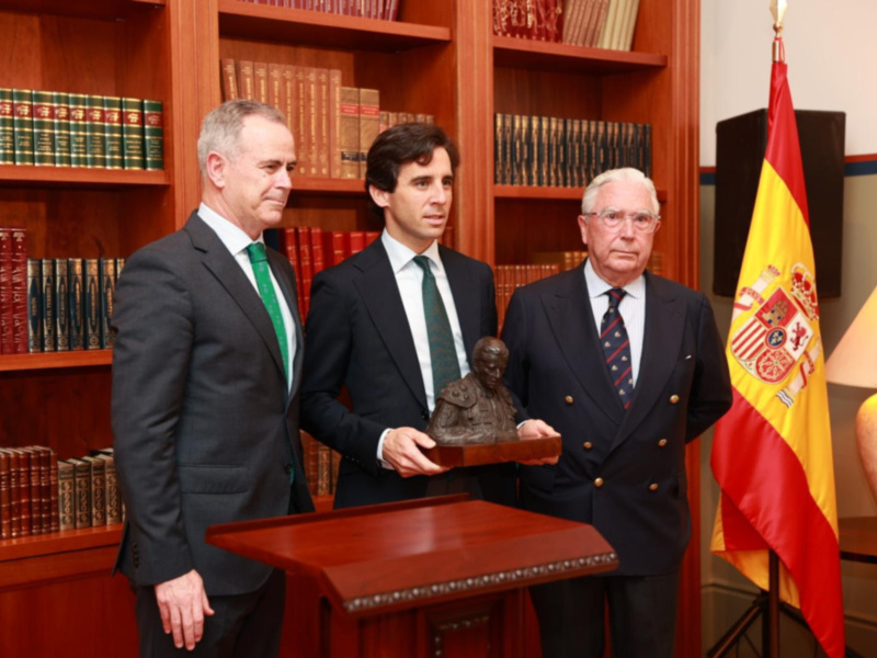 Tres hombres en trajes formales posan con un busto en un entorno académico con libros y bandera española.