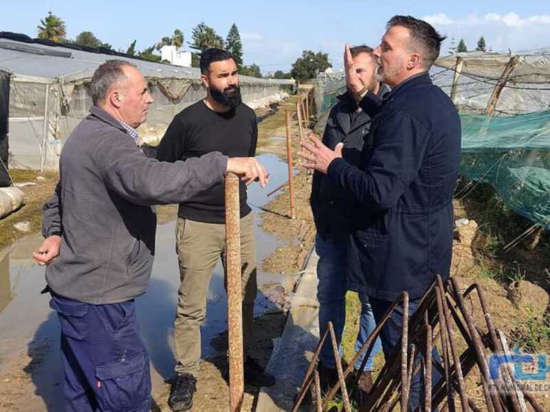 Tres personas conversan en un entorno agrícola, posiblemente en un huerto o invernadero, con estructuras de metal y cubiertas de plástico en el fondo.