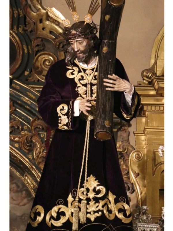 Imagen de un Cristo de la Pasión en trono, vestido con un manto negro bordado con oro y portando una cruz, en un entorno religioso con decoración dorada.