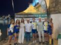 Equipo de remo celebrando una victoria en el Campeonato Andaluz de Remo, posando frente a un stand de la Federación Andaluza de Remo, con el río y la torre de la Giralda al fondo en la noche de Sevilla.