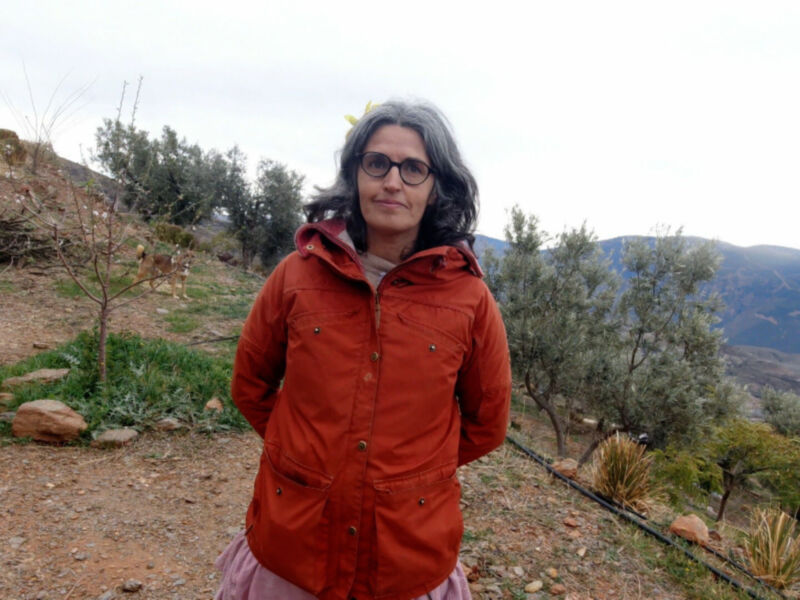 A una persona con cabello gris y gafas, vestida con una chaqueta naranja. El fondo es un paisaje desértico con pequeños árboles y montañas en el horizonte. La persona parece estar en un camino de tierra, con rocas y vegetación dispersas alrededor.