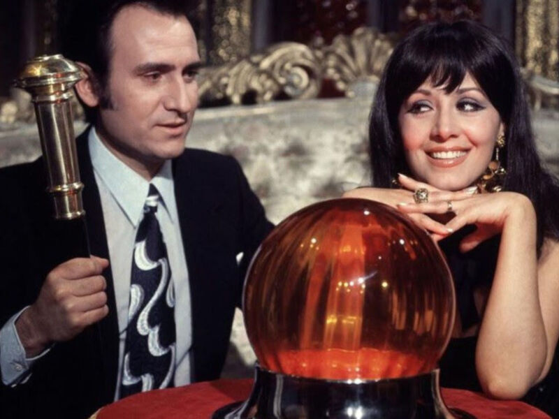 Un hombre y una mujer están sentados en un sofá de terciopelo, con el hombre sosteniendo una bola de cristal roja y brillante. La mujer sonríe mientras mira a la cámara, ambos parecen estar en un entorno elegante y sofisticado.