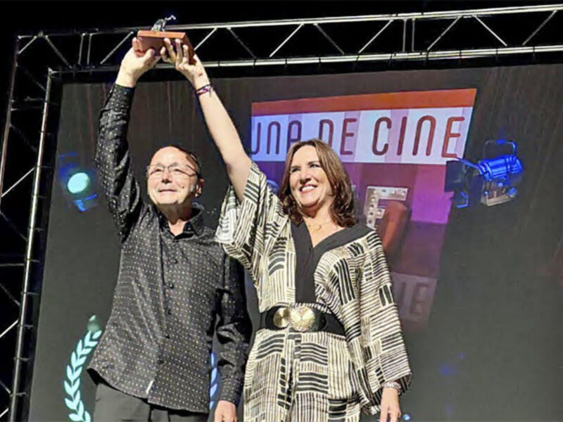 Dos personas celebran con un premio en una gala de cine, frente a un escenario con la bandera "UNA DE CINE".