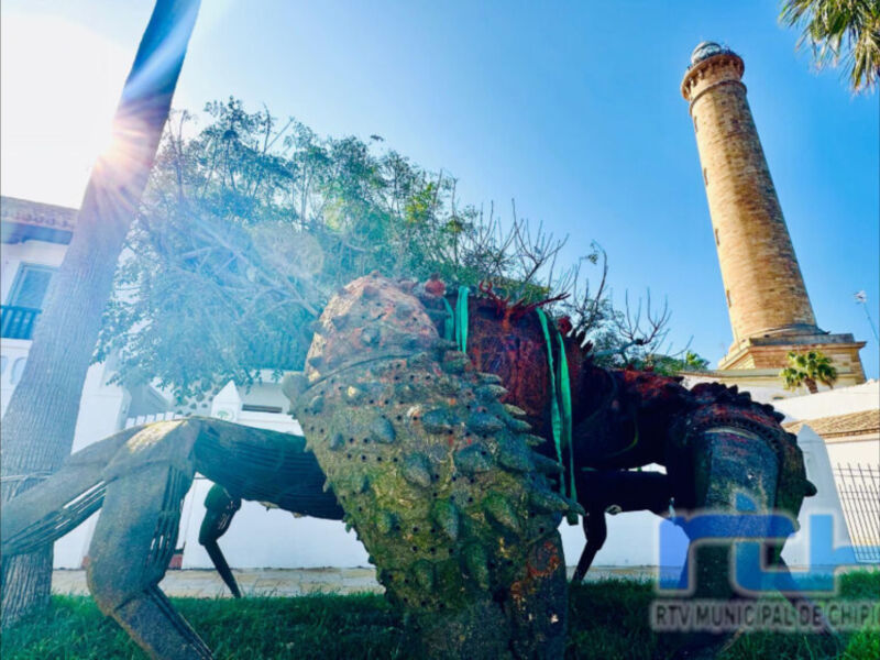 Un dragón de metal oxidado con detalles verdes y azules, enmarcando una torre de ladrillo amarillo. El cielo azul claro y la vegetación cercana completan el paisaje urbano.