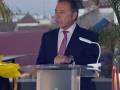 Un hombre en traje negro y corbata rosa se encuentra detrás de un podium con el logotipo de la Junta de Andalucía. La imagen muestra una vista de un edificio con tejas y cielo claro en el fondo, probablemente durante la tarde.