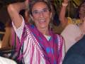 Una mujer sonríe ampliamente mientras levanta su mano izquierda, con una pañuelo de color rosa y azul con puntos en forma de corazón. La imagen muestra a varias personas en un ambiente nocturno, posiblemente una fiesta o evento.