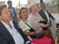 Tres personas sentadas en un evento, con una mujer en el centro y dos mujeres a sus lados. La imagen muestra detalles de la vestimenta y una mano con un bolso negro. En el fondo, se puede ver una estructura arquitectónica y un cielo despejado.
