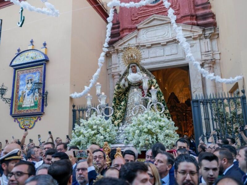 Fotografía de una procesión religiosa con una imagen de la Virgen María en el centro, rodeada por una gran corona de flores y un grupo de personas celebrando. La imagen se realiza en frente de una iglesia con detalles arquitectónicos y decoración.