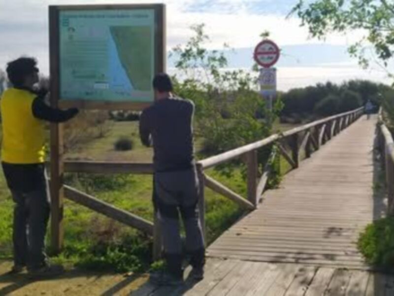 Dos personas observan una pantalla digital en un sendero de madera, con un cartel y un signo en el fondo.