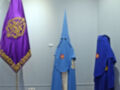 Imagen de dos figuras vestidas con capas azules y un escudo rojo, junto a una bandera morada con símbolos dorados. El fondo es un muro de color claro con una toma de corriente y un ventilador.